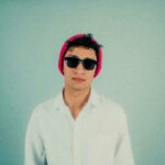 Fashionable young man wearing sunglasses and a pink beanie, set against a minimalist pastel background.